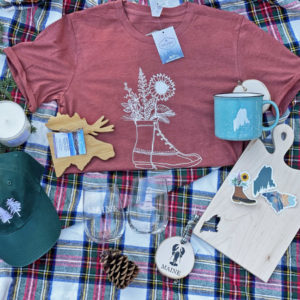 A tshirt with a llbean boot bouquet, serving board with state of Maine cutout, mug, etched glassware, hat with two trees on it, ornaments and more laying on plaid blanket