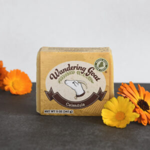 Wandering Goat Calendula Bar Soap. A bar of wrapped soap with a goat logo sits on a piece of grey slate with fresh orange and yellow calendula flowers to the right of the soap and in the background.