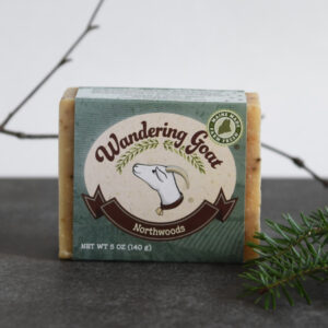A bar of soap with a green label and goat sits on a piece of grey slate. There is a fresh sprig of spruce in the foreground and a winter tree branch in the background.